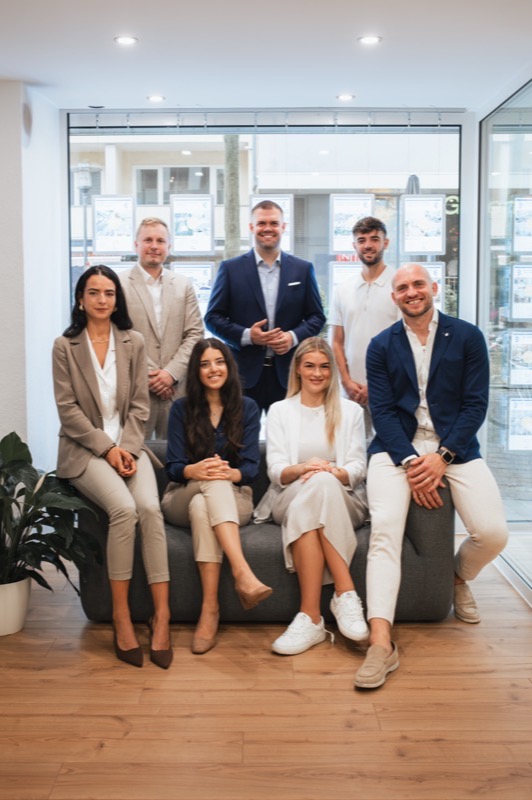 Teamfoto: acht Mitarbeiterinnen und Mitarbeiter eines Immobilienbüros auf und hinter einem Sofa vor dem Schaufenster