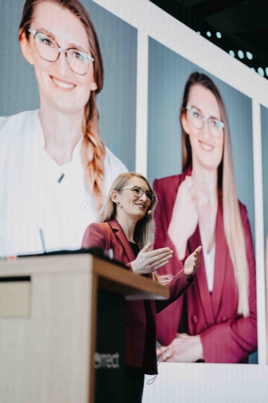 Rednerin im roten Blazer spricht am Pult vor großen Portraits auf der Leinwand