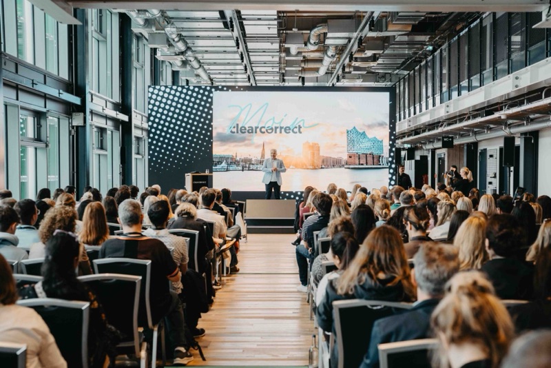 Redner auf der Bühne einer Firmenkonferenz vor vollem Saal in einer modernen Industriehalle