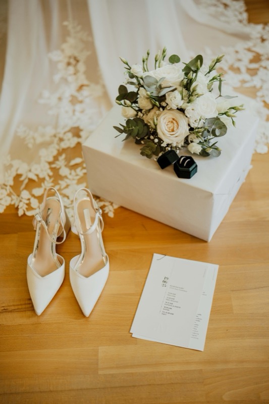 Brautschuhe, Brautstrauß und Ringbox auf Holzboden vor dem Brautkleid