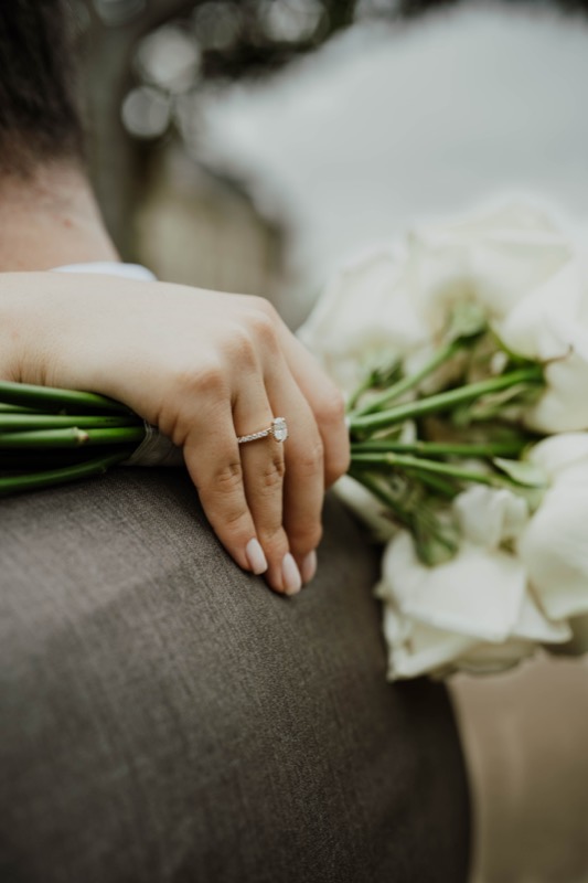 Hand der Braut mit Verlobungsring auf der Schulter des Bräutigams, daneben weiße Calla