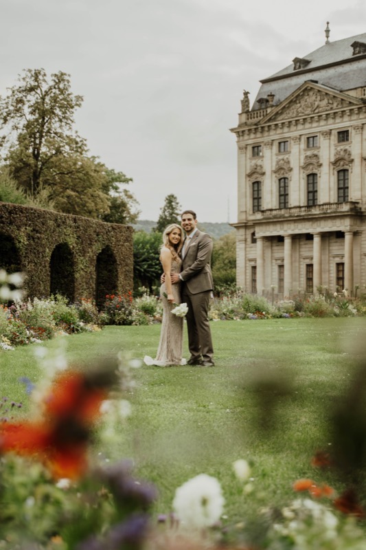 Brautpaar weit entfernt im Schlossgarten, im Vordergrund unscharfe Blumen