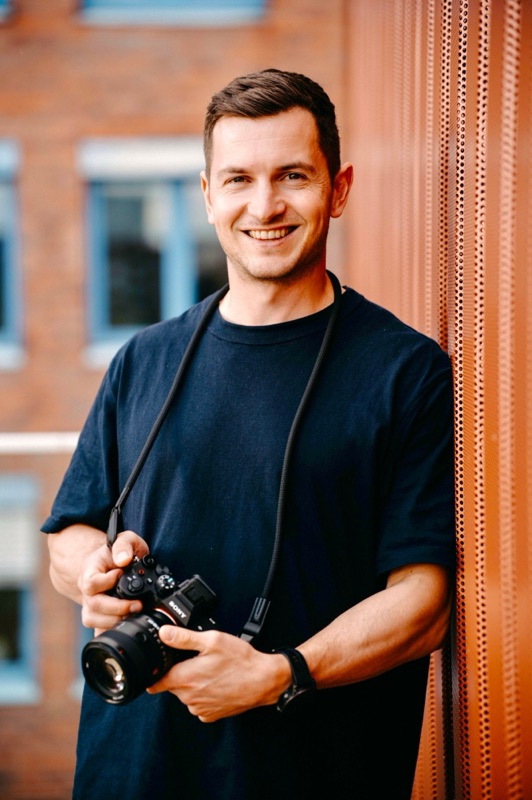 Fotograf Ken Stüker lächelt mit Kamera in der Hand vor einer Backsteinfassade